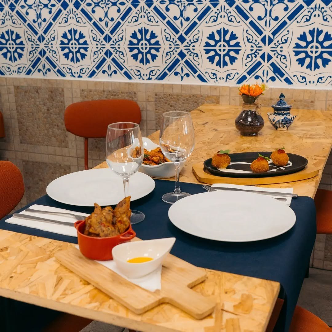 Mesa posta com toalha azul, pratos, copos e petiscos, junto a uma parede de azulejos azuis e brancos